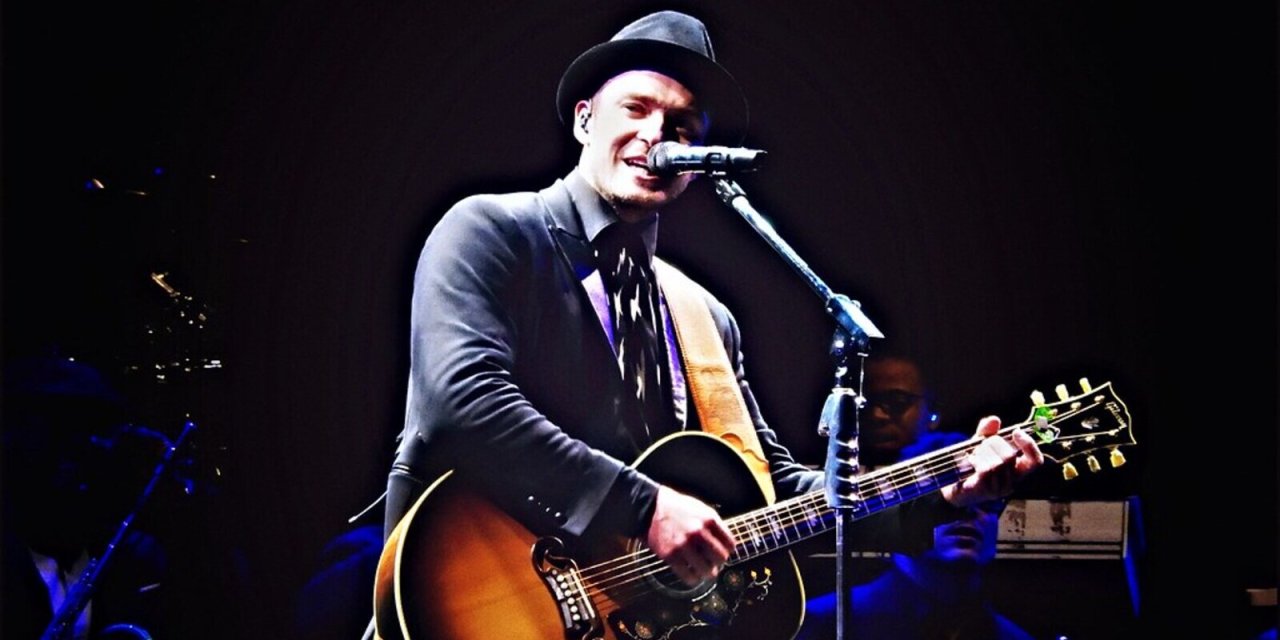 Justin Timberlake wearing a hat holding a guitar singing on stage 