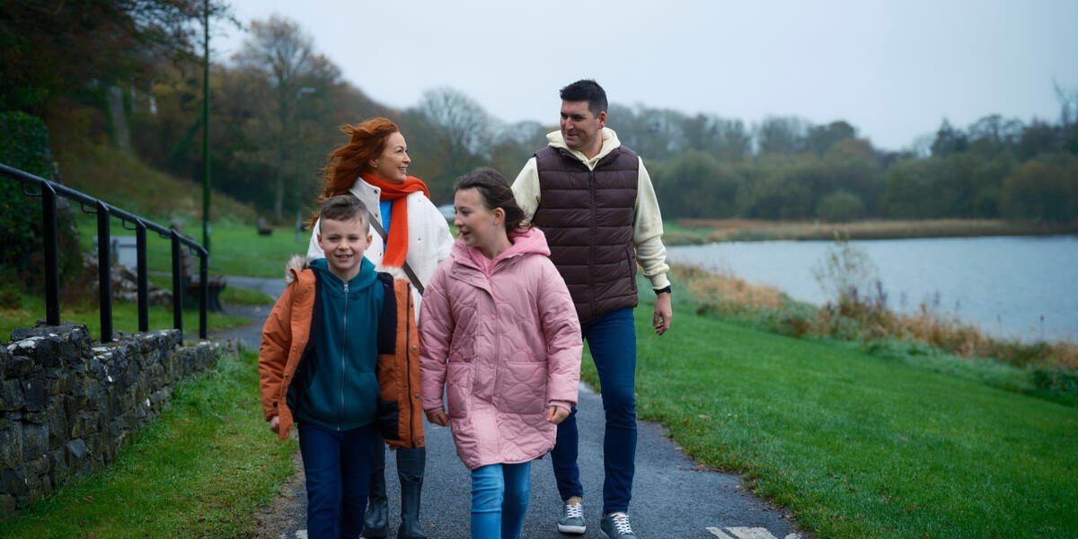A happy family of four walks along a pathway beside a lake in autumn