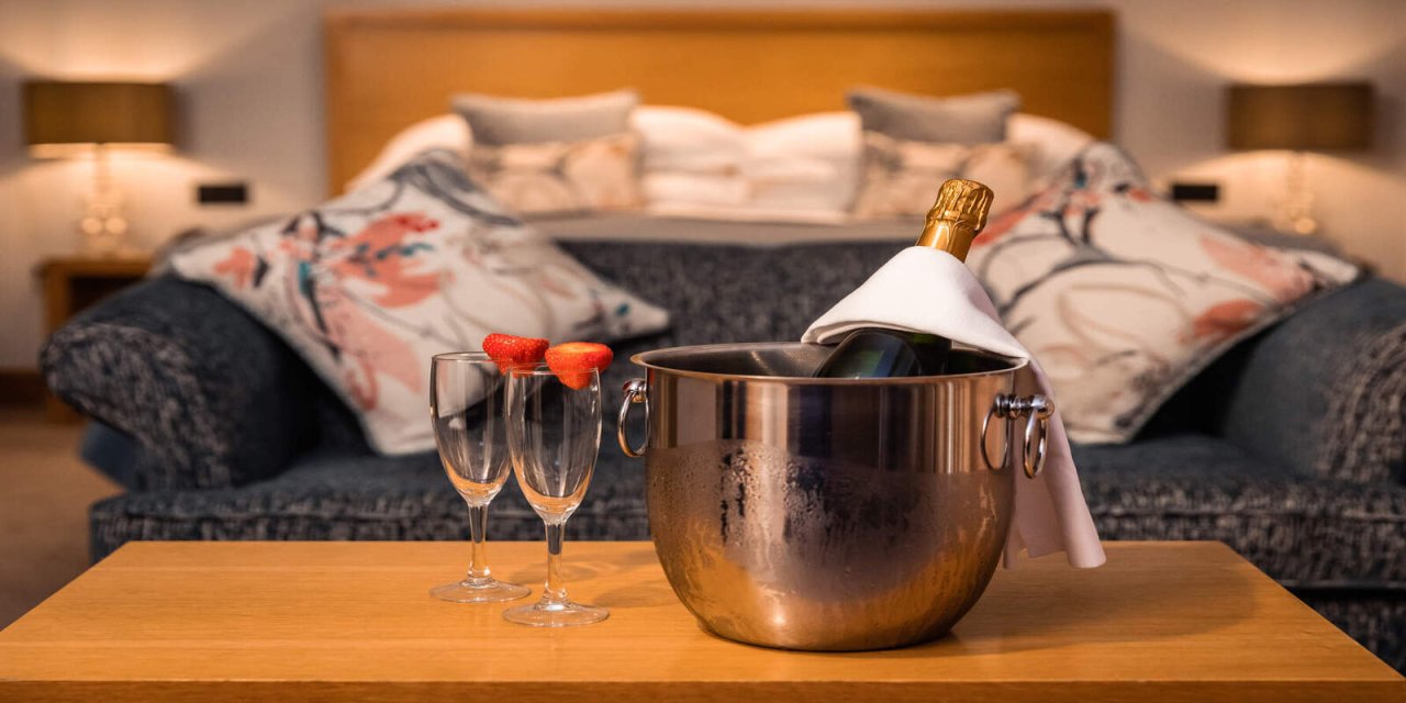 Hotel room with silver ice bucket and chilled glasses with strawberries, Cold bottle of prosecco sitting on a table in front of a cozy couch 