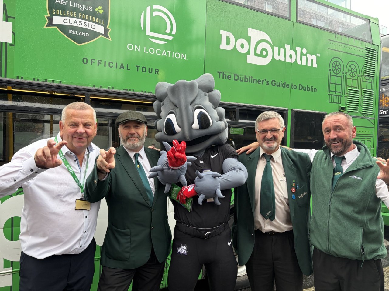 3 dodublin drivers wearing green jackets, one man in white shirt and football mascot in black t shirt smiling infront of green dodublin bus