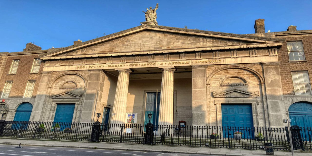 Old stone church building with statue of a figure on the roof. The roof comes to a triangular point. There are two paneled columns in the middle of the building, helping holding it up. There are 4 bright blue doors - 2 are cornered off and the other two have oval tops. There are stairs leading up to the building which isprotected back a black fence.