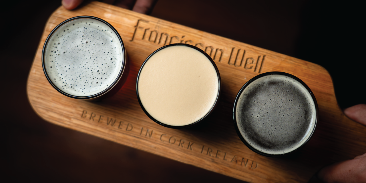 3 stout beers with foam on top sitting on a board that is placed on a table in a dark lit room. 