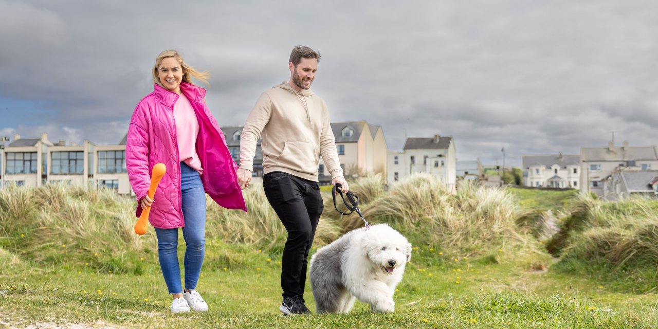 A smiling woman in a bright pink jacket and a man in a beige hoodie walk hand in hand with a fluffy white and gray dog through grassy coastal dunes under a cloudy sky.