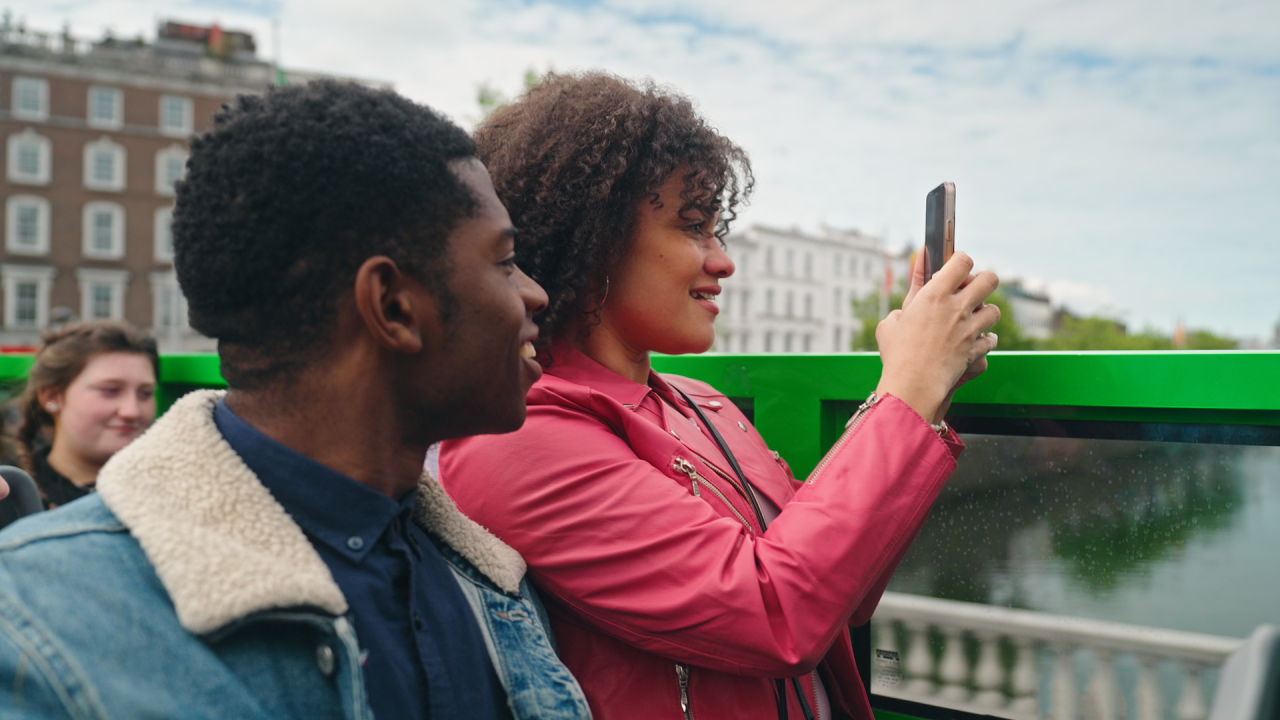 a woman and a man sitting beside each other on the open top tour bus and the woman is taking a picture using her phone