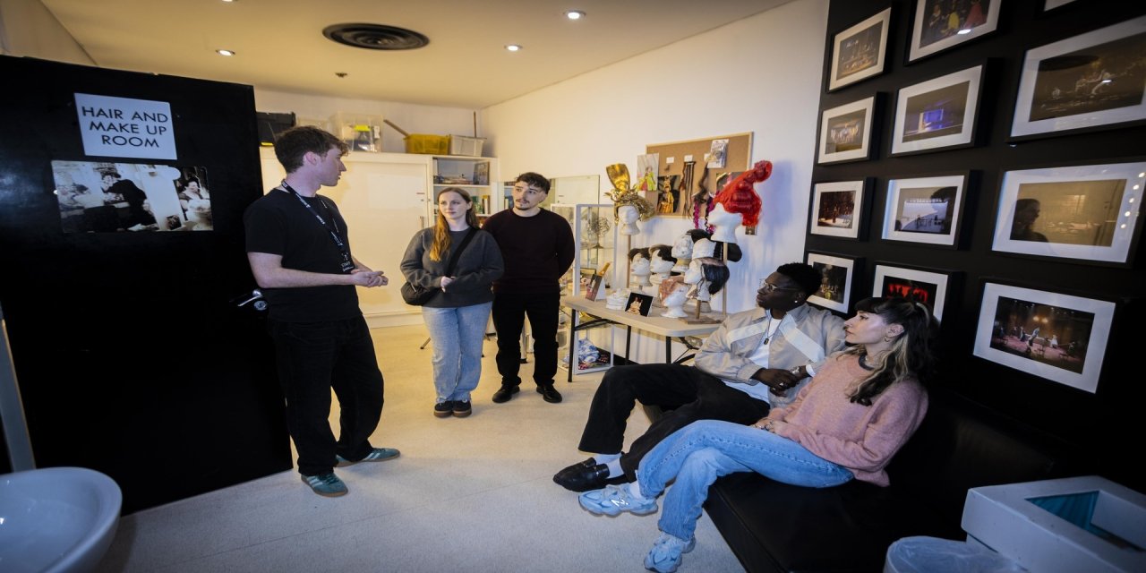 Backstage room black walls with pictures hanging up. People standing and listening to guide