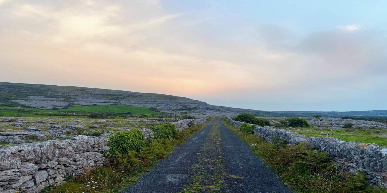 Ombre sky with large cloud, sun setting with walking path in the middle of the photo. Short stone walls on either side of the path with greenery in the filed beyond them. 
