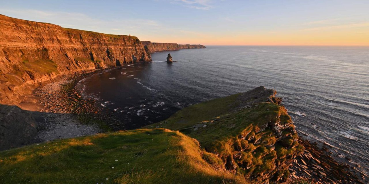 towering brown sea cliffs overlooking the dark navy ocean water with ripples in ocean. Orange and blue sun set in the background with small waves crashing against the rocks