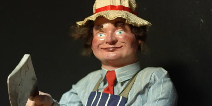 museum-of-curiosities-doll old plastic doll holding an axe. Wearing white hat with red ribbon, a red rie, light blue striped shirt and dark blue stripe gown