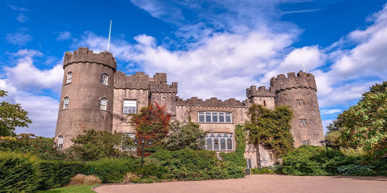 Tall round stone castle with tall bushes and green trees surrounding it. Sand long driveaway