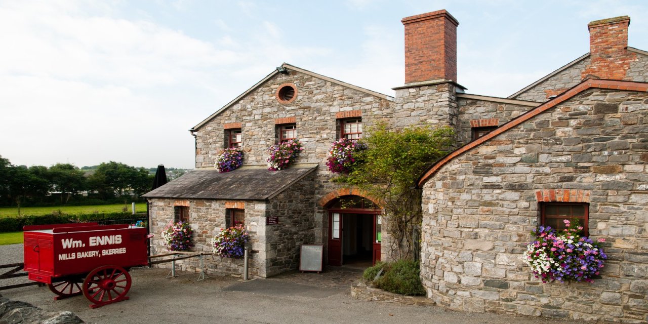 Grey bricked mill with red wheel barrow on the left foreground. Purple, white and pink flowers hanging from the windows of the mills. Rd brick chimney.   